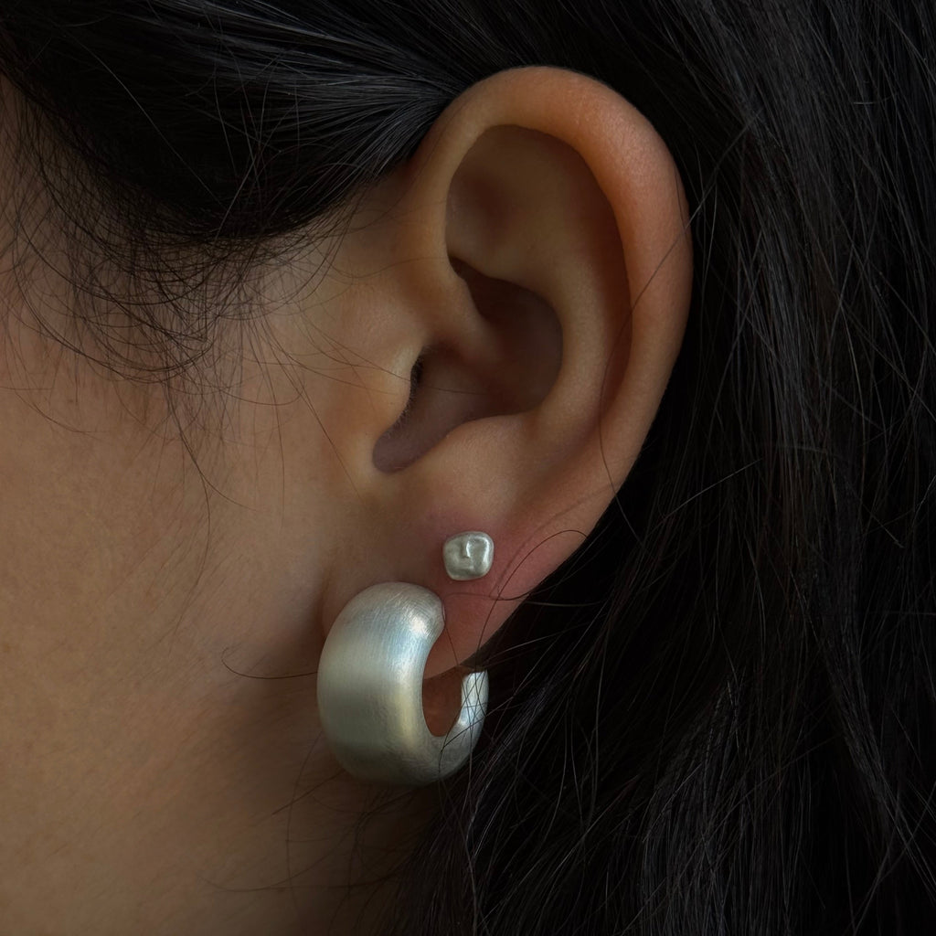 Close-up of an ear wearing a silver hoop earring with a small stud BY EM JONO